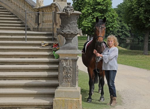 Marion Becker steht neben einem Pferd vor einer Freitreppe in Moritzburg.