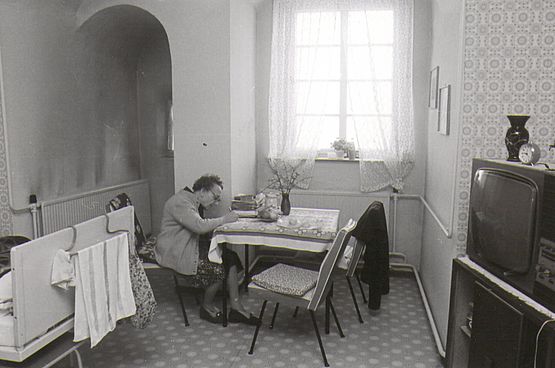 Die Fotografie zeigt einen Einblick in den Wohnraum des Altersheimes. Eine alte Dame sitzt an einem Tisch am Fenster und schreibt.