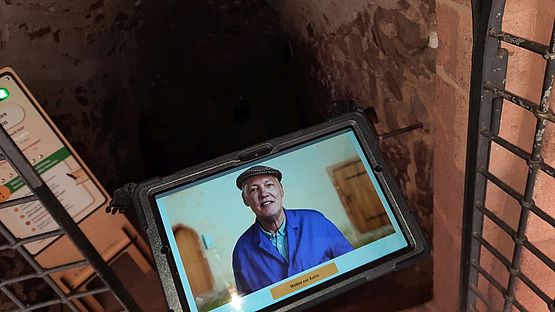 Eine Hand hält ein Tablet, auf dem Hausmeister Herbert zu sehen ist. Im Hintergrund sieht man einen dunklen Treppenabgang, der in einen Keller führt.